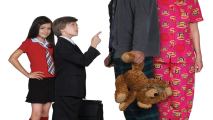 A young girl and boy, dressed in business attire, are standing in front of two adults dressed like kids, in pyjamas and holding a teddy bear.