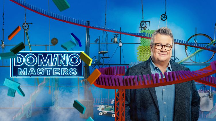 A man wearing glasses and a jacket standing in front of a blue wall, with a logo on his left side that says Domino Masters.
