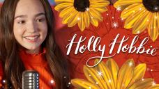 A close-up of a teenage girl in front of a microphone, next to a logo that says Holly Hobbie.